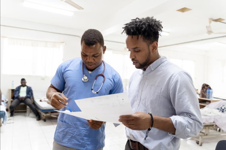 A Haitian clinician in light blue scrubs reviews a series of paper with a colleague, who stands to his right.