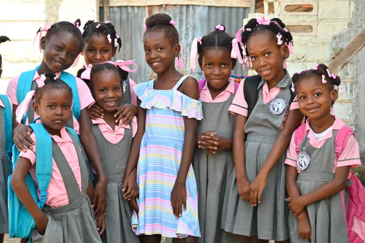 A girl in blue & white dress smiles shyly and happily. She is surrounded by 7 other smiling girls in gray & pink school uniforms.