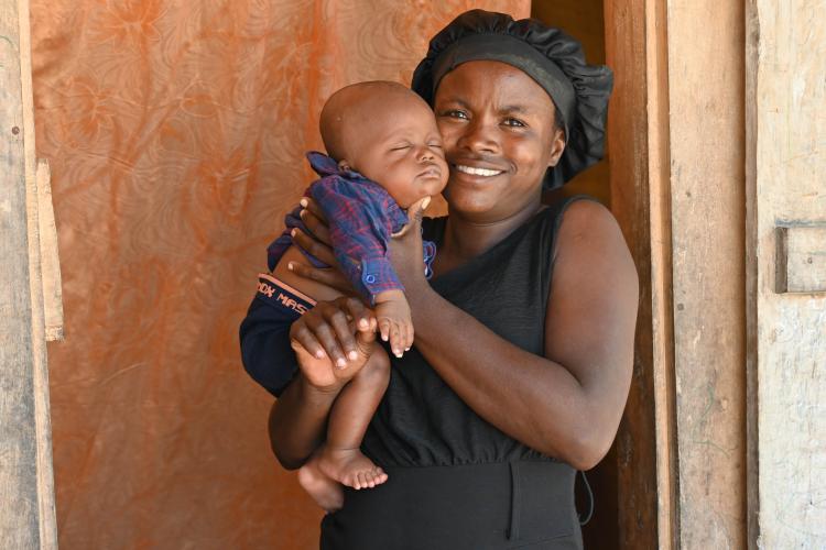 A Haitian woman stands in a wooden doorway. She holds her infant son in her hands, held up near her face. He wears blue shorts and a plaid button down-shirt. He is sleeping. The woman wears a black hair bonnet and a sleeveless black dress. She has a bright smile.