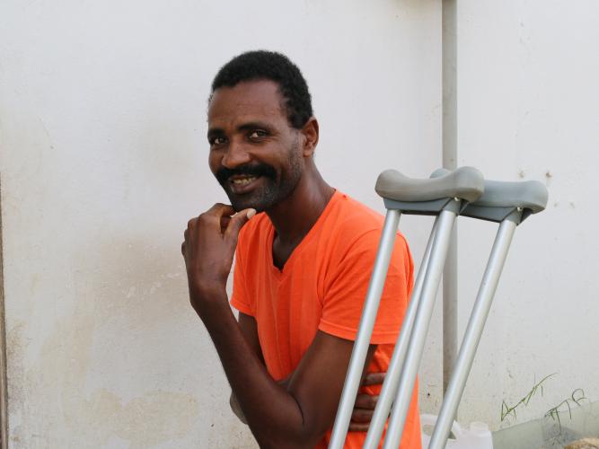 A Haitian man smiles playfully with his hand to his chin. A pair of crutches lean next to him.