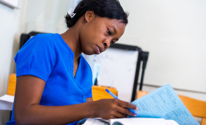 Sitting at a table, a Haitian nurse in bright blue scrubs studies a document she is holding.