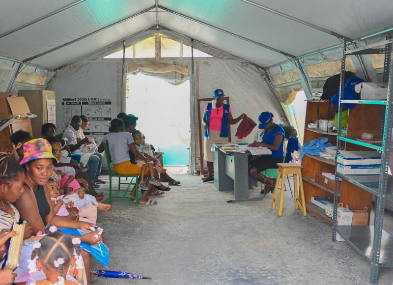 Interior view of a large tent. On the left, the family in the previous picture waits alongside other parents and children. On the right, there are health workers and shelves of supplies.
