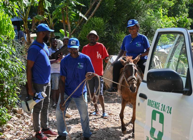 With a white ambulance to one side, a man in blue T-shirt and cap leads a mule loaded with supplies and carrying a woman in blue T-shirt. Local residents look on, smiling.
