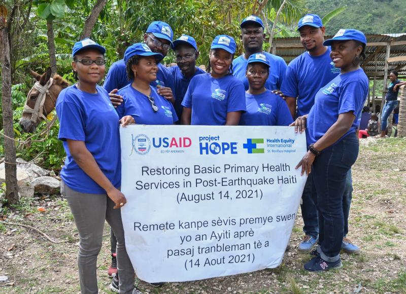 9 Haitians in blue HEI T-shirts and caps pose, smiling, with a sign that says, “Restoring Basic Primary Health Services in Post-Earthquake Haiti.”
