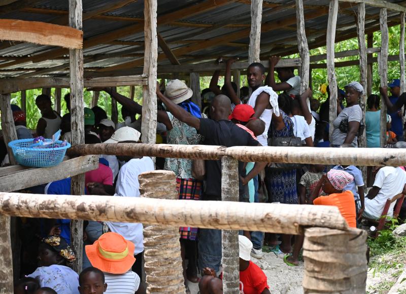 Alt: A large number of Haitian men, women, and children wait in the shade of a corrugated tin roof held up by rough-hewn wooden posts.