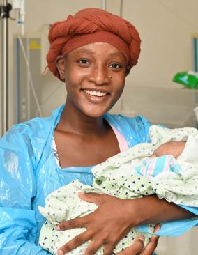 A young Haitian woman wearing a orange-brown head wrap and blue hospital gown smiles happily. She holds a small infant in her arms.