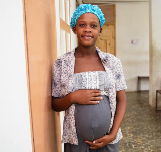 A pregnant Haitian woman stands in the courtyard hallway at St. Boniface Hospital. She wears a grey dress with a white lace top, a open flower-printed shirt, and a bright blue hair bonnet. She smiles at the camera with her hands on the top and bottom of her pregnant stomach.