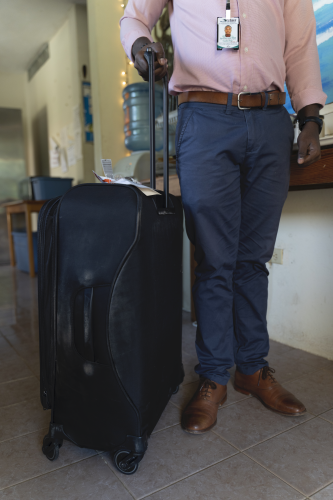 A tall Haitian man stands next to a black suitcase. The man wears business attire.
