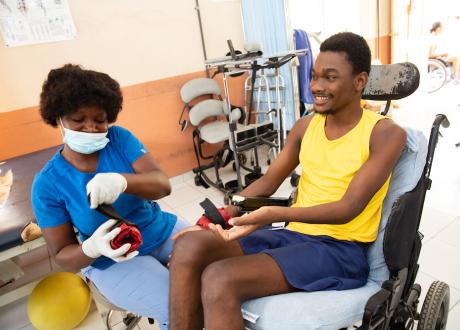 A Haitian man sits in a wheelchair. He wears a yellow tank top and blue shorts. He holds a wrist weight in his cupped hands. A Haitian nurse sits next to him, she is wearing blue scrubs and is adjusting one of the wrist weights. They smile and laugh to each other.