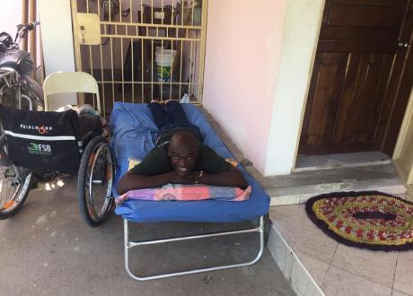 A Haitian man reclines on his stomach on a blue cot. His wheelchair sits next to the bed.