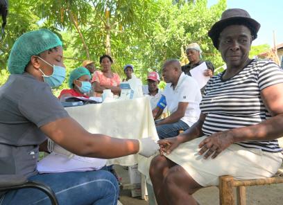 Under a tree, a woman wearing a face mask and gloves checks an older woman’s vital signs. Six more adults are in the background.