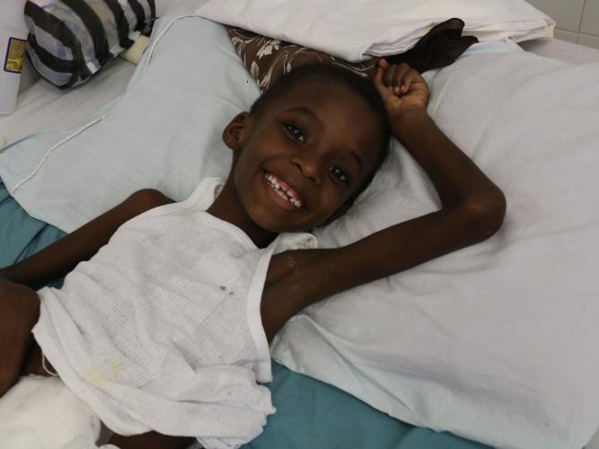 A young Haitian boy lays on a hospital bed in a white tank top. He smiles and holds his arm above his head.