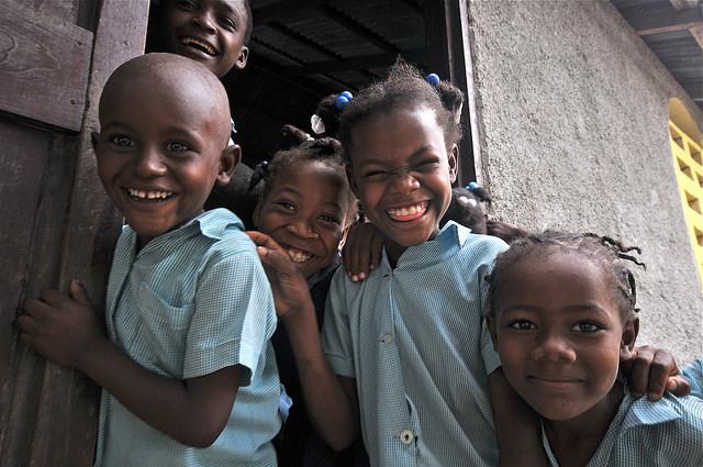 A group of five Haitian school children laugh and smile. They are wearing blue checked shirts.