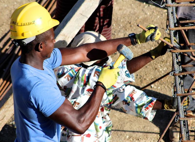 Two Haitian construction workers in hardhats work on a rebar support structure using hammers.