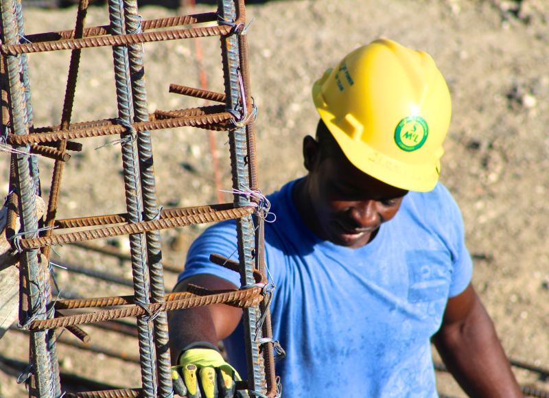 A construction worker in a blue t-shirt and yellow hardhat holds onto a rectangular rebar structure at a construction site.
