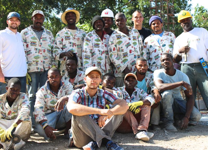 A team of construction workers pose for a group photo outside. They are arranged in two rows: several men stand in the back and more sit cross-legged in the front.