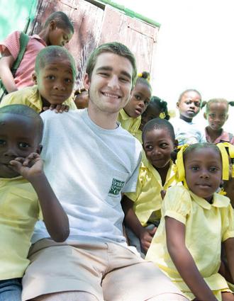 A young white man sits with a crowd of young Haitian schoolchildren wearing school uniforms.